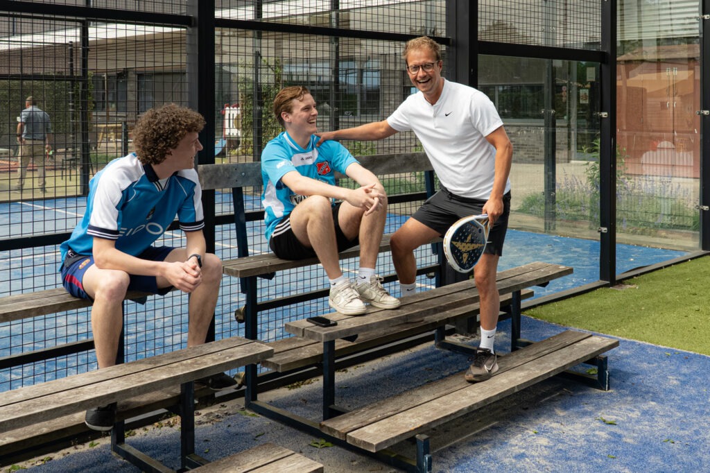 Drie leden van het team zitten gezellig op het toeschouwers bankje met elkaar te lachen tijdens het padellen.